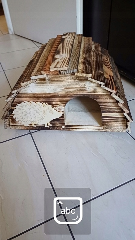Drewniany domek dla jeża z wyciętym otworem i drewnianym jeżem z boku.